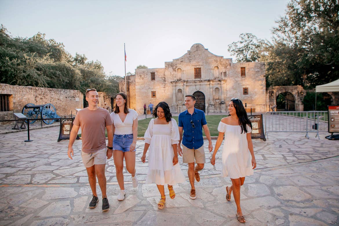 Two men and three women walk outdoors near the Alamo during the summer.