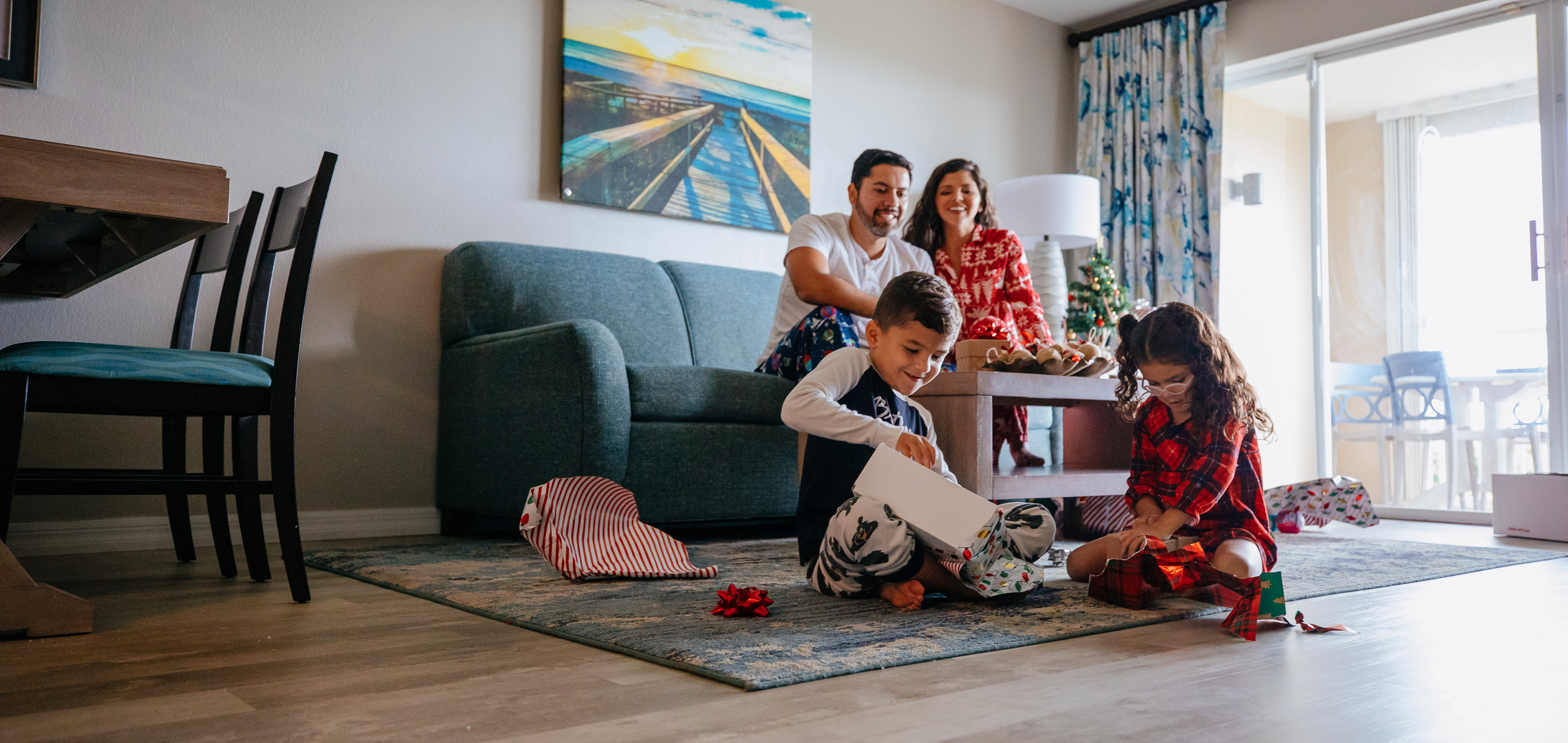 A man, young boy and woman watch a young girl unwrap a present in a living room while all wearing pajamas.
