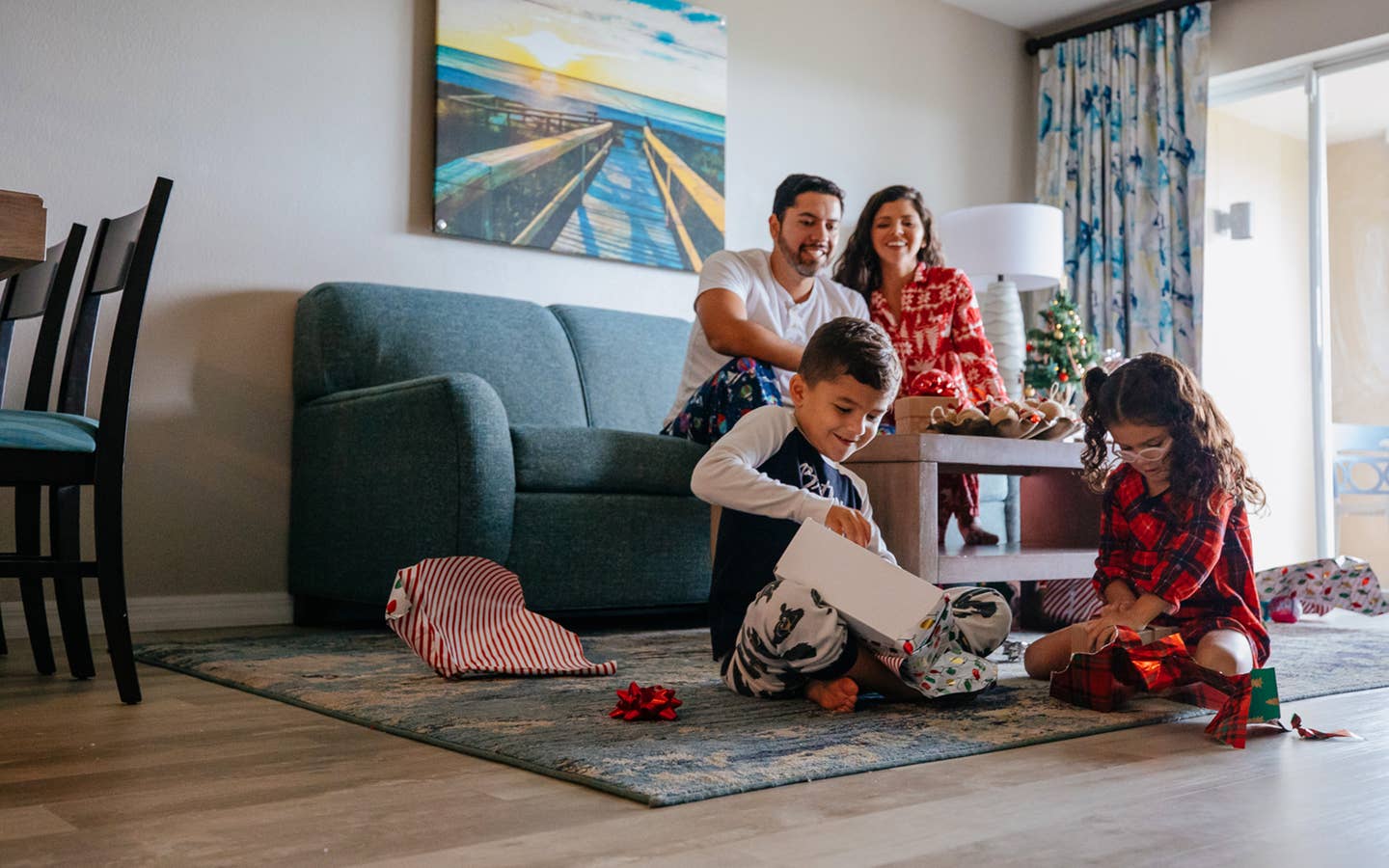 A man, young boy and woman watch a young girl unwrap a present in a living room while all wearing pajamas.