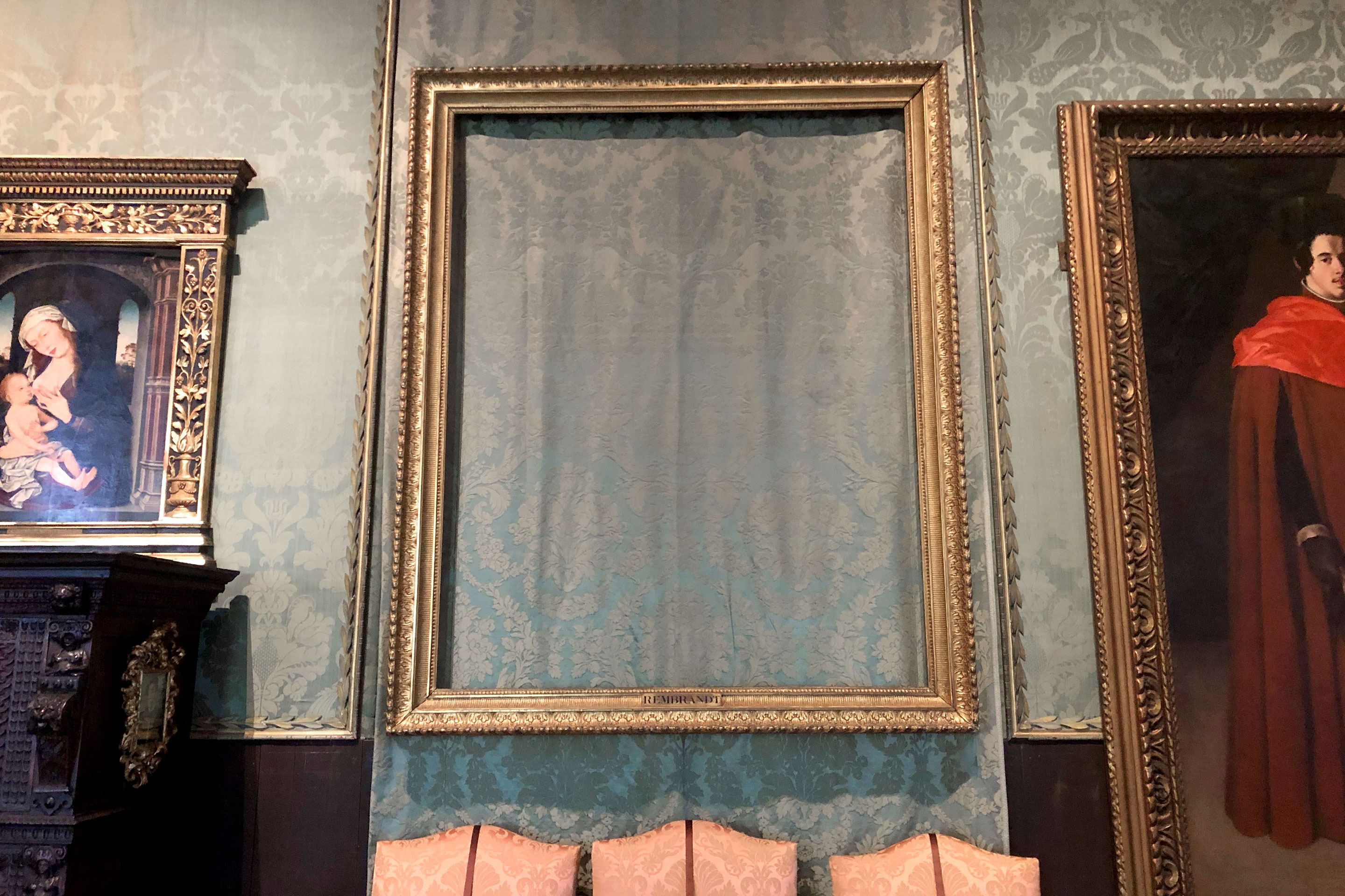An empty, gold frame hangs with a plaque reading 'Rembrandt' surrounded by paintings on a wall.