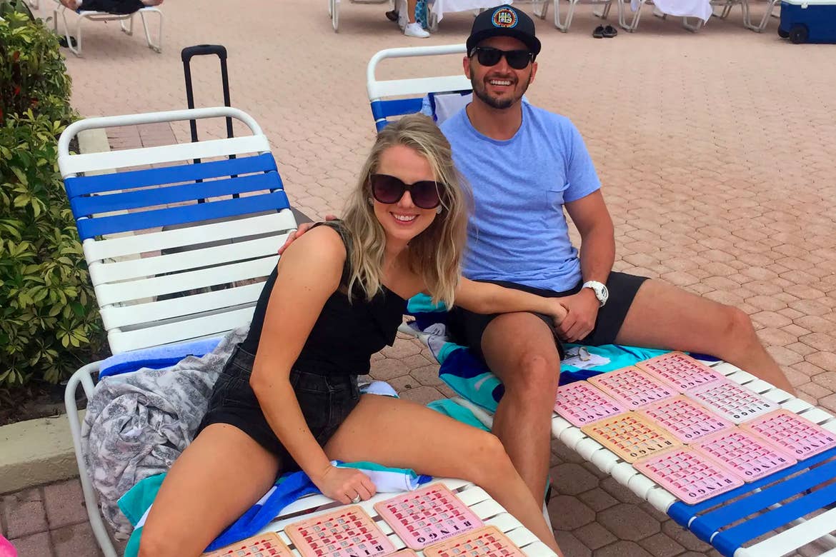 A caucasian woman wearing a black blouse and shorts (left) and Latinx male wearing a blue t-shirt, black shorts and a black hat (right) sit on two lounge chairs with numerous Bingo cards.