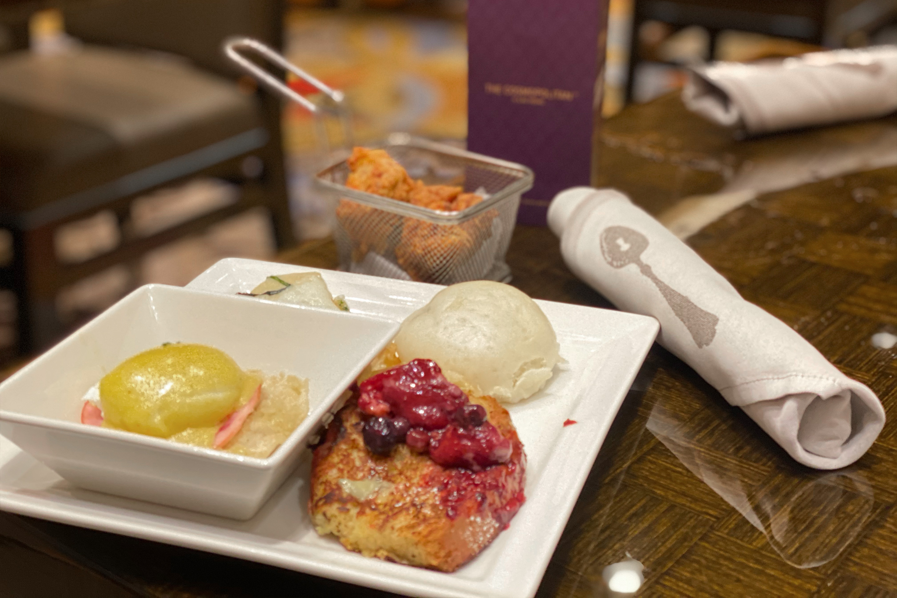 A white square plate containing bread pudding and a white square bowl containing eggs Benedict are placed on a wooden dining table near silverware wrapped in a white napkin.