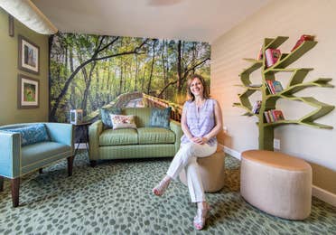 Woman sitting in a room with forrest wallpaper and books on a shelf.