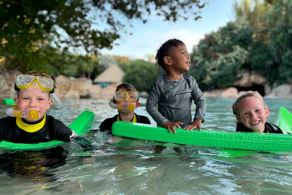 Three children in wetsuits and snorkel gear swim near a young boy in a grey wetsuit holding green floaties in an outdoor water area.