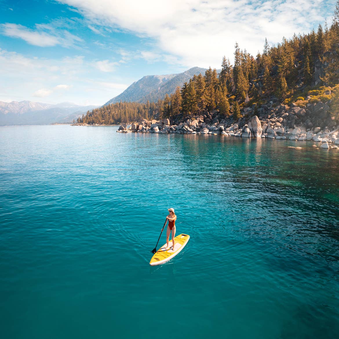 A caucasian woman wears a red one-piece swimsuit and straw sunhat as she stands on a yellow stand up paddle-board in a lake surrounded by sunlight, cloudy blue sky and pine trees.