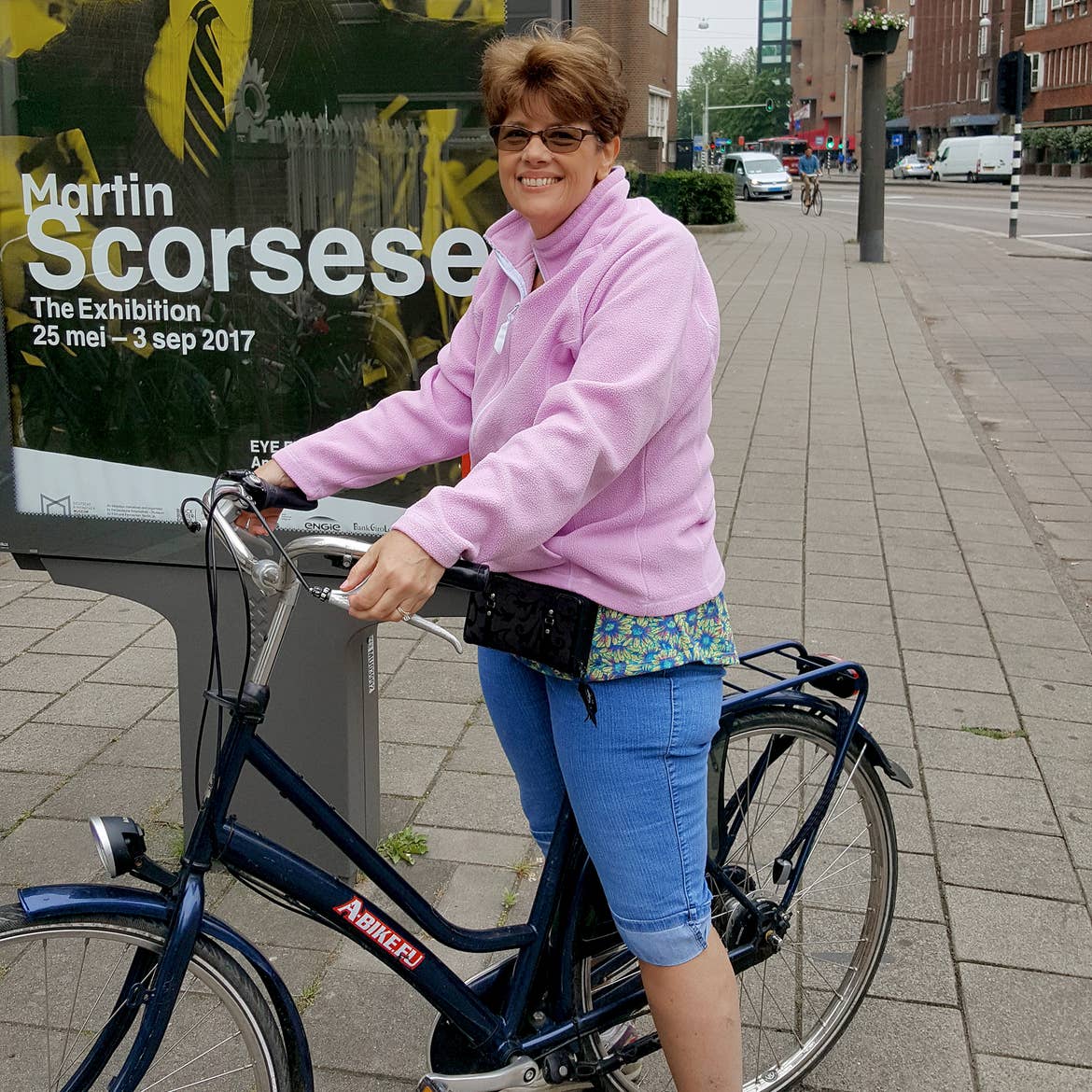 A caucasian woman wearing a pink fleece sweater and jeans sits atop a bicycle on the streets of Amsterdam.