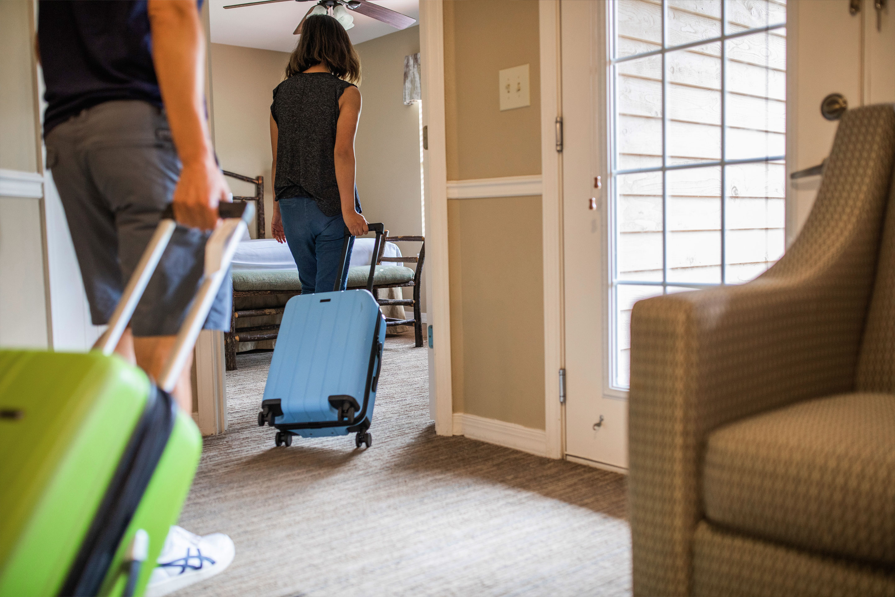 A woman and man bring green and blue suitcases into a villa bedroom.
