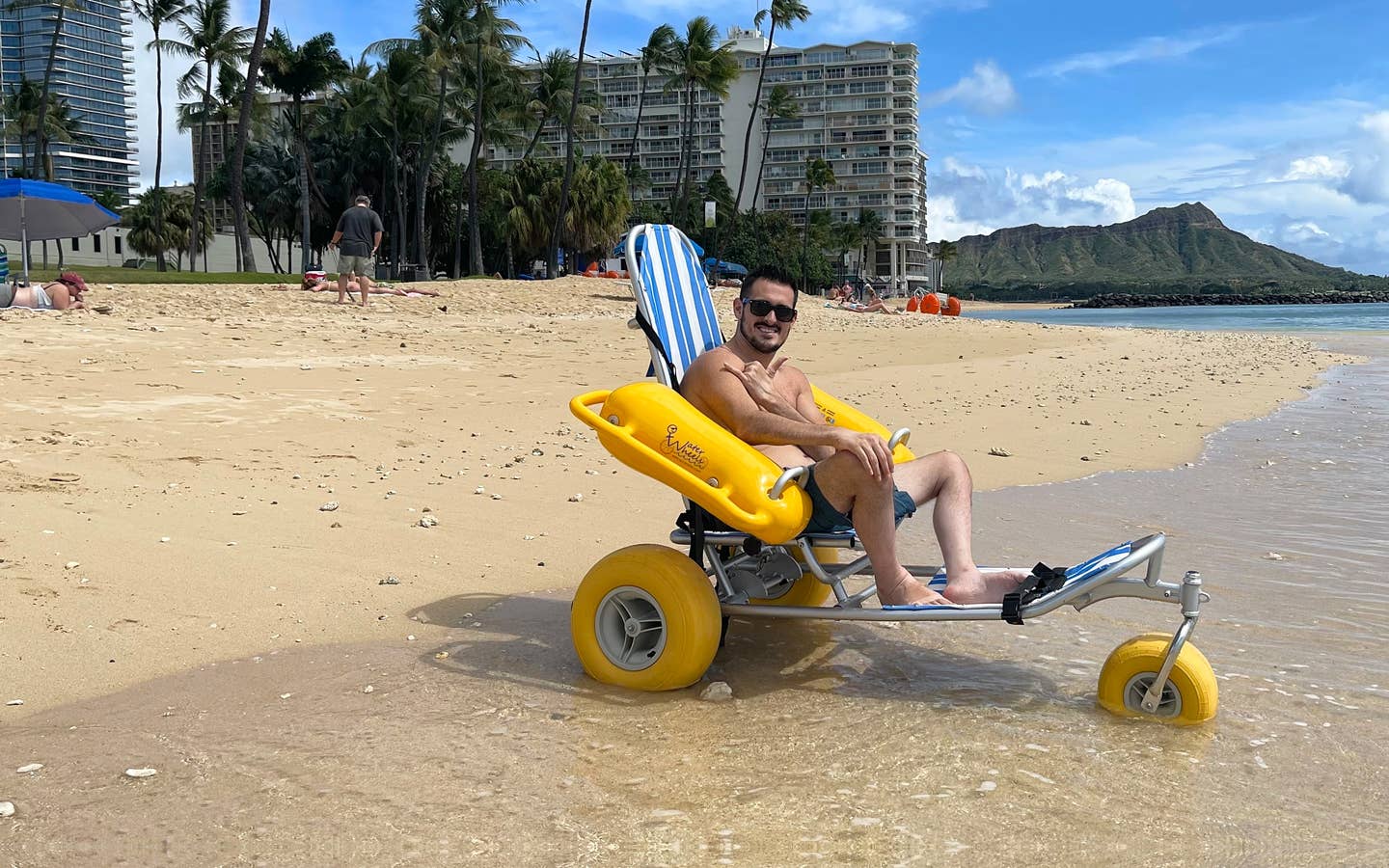 Featured Contributor, Danny Pitaluga, wears sunglasses and swim trunks while sitting in a yellow beach wheelchair on the sand as waves roll in.
