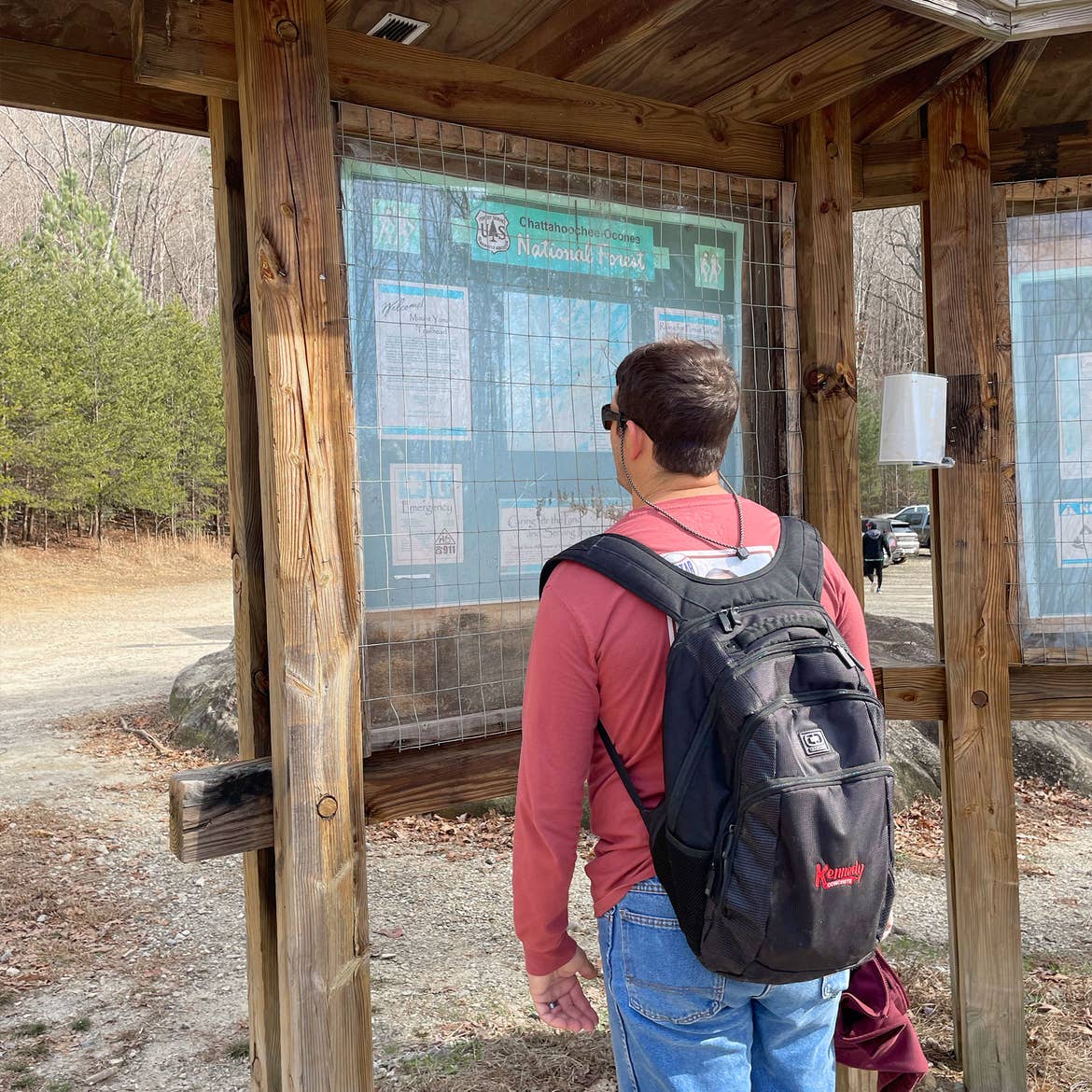 A man in sunglasses, a red long sleeve shirt, denim pants and black backpack reads trailhead signage.