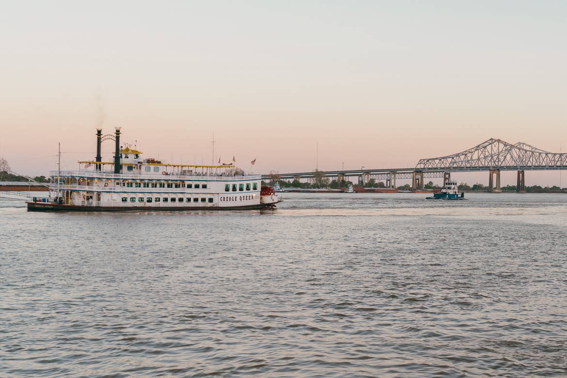 A riverboat named the 'Creole Queen' floats along the Mississippi River in front of the Crescent City Connection bridge under a sunset sky.