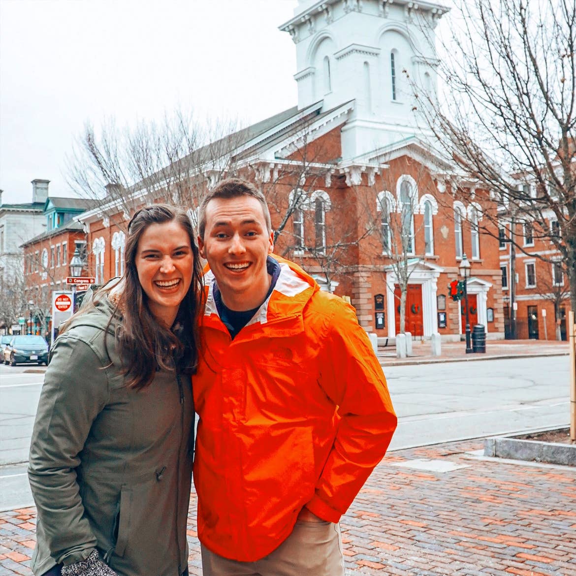 A woman in a green jacket and a man in an orange jacket pose on a snow-covered street near a historical brick building.