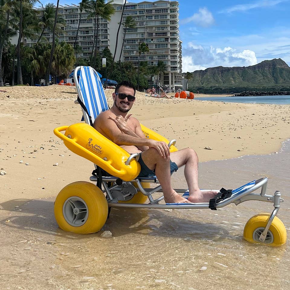 Featured Contributor, Danny Pitaluga, wears sunglasses and swim trunks while sitting in a yellow beach wheelchair on the sand as waves roll in.