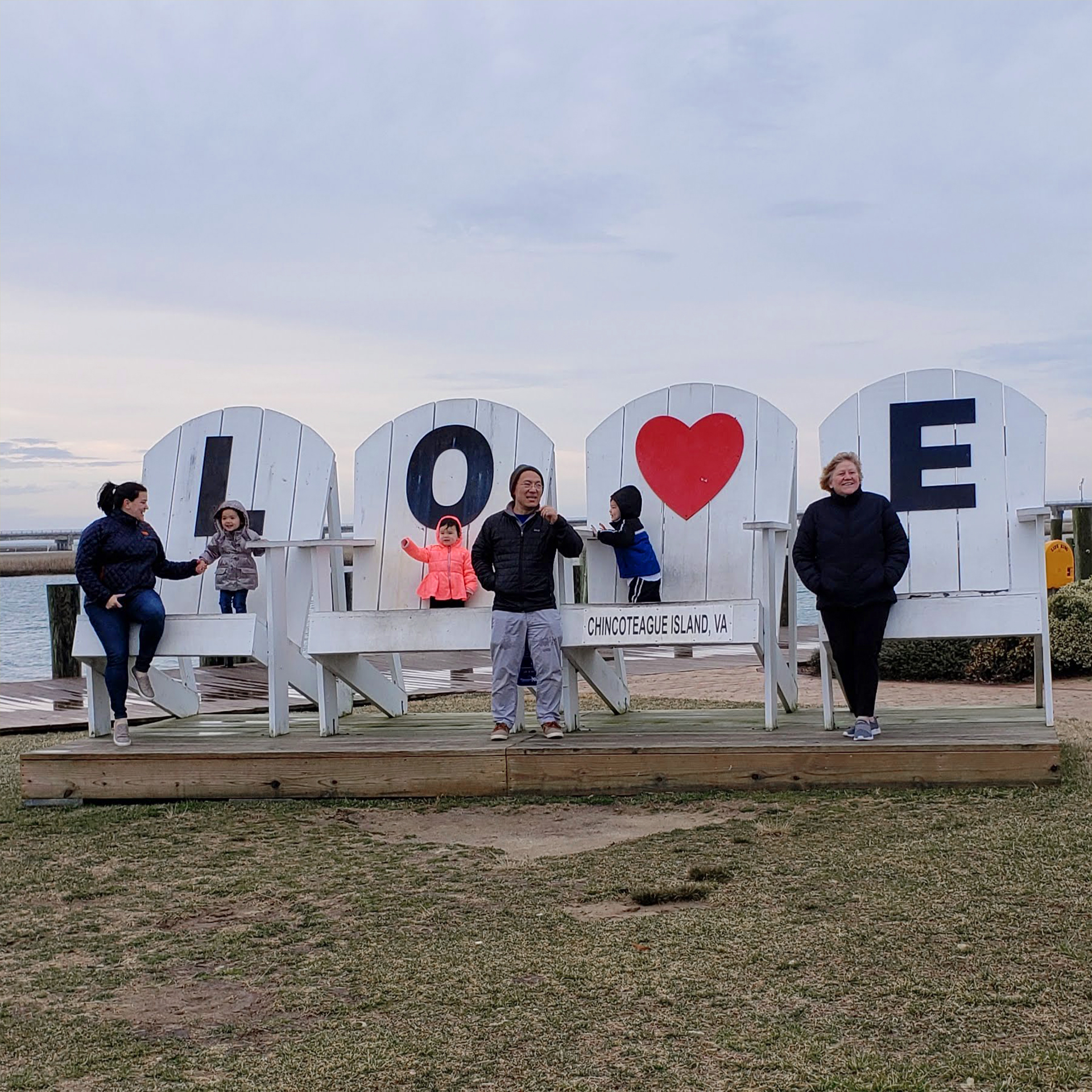 Three Asian Pacific Islander toddlers (left to right: Two girls and a boy) stand on large adirondack chair outdoors wearing winter jackets near three adults.