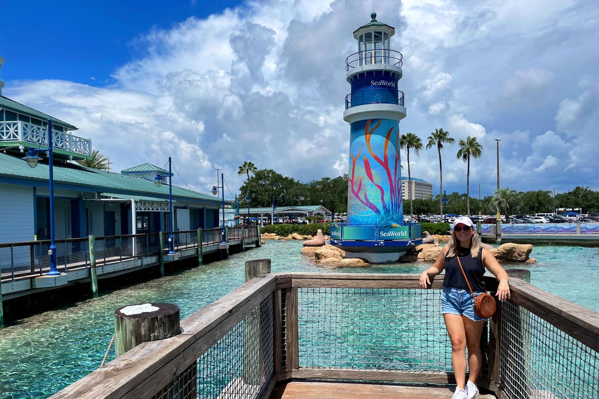 A Caucasian female wearing a pink baseball cap and black tank top stands near the lighthouse in SeaWorld Orlando.