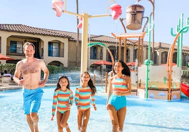 (From left to right) A man, two young girls and a woman in matching swimwear sun away from an outdoor splash pad area near a building exterior under palm trees and a blue sky.