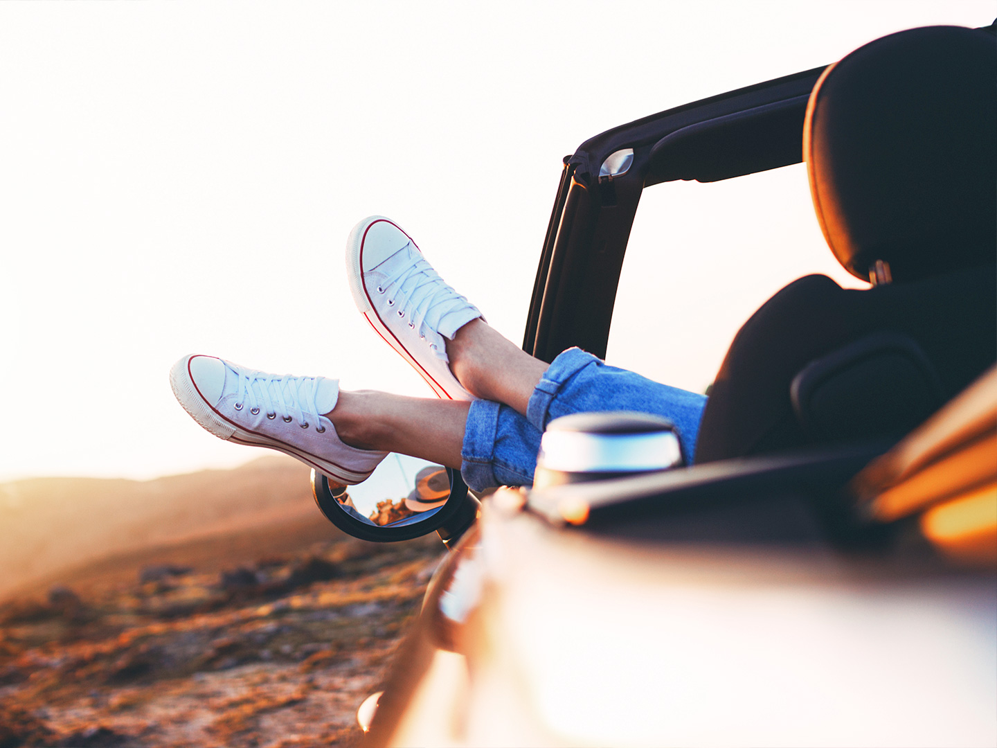 A woman wearing white sneakers sticks her feet out of the convertible car near a beachfront as the sun sets.