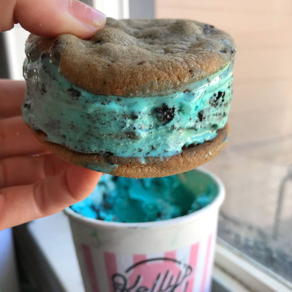 A woman holds a blue ice cream, chocolate chip cookie sandwich over a pint of ice cream.
