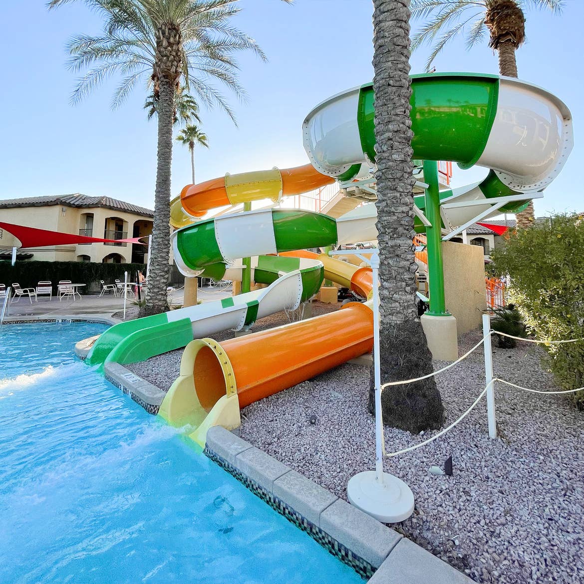 Two swirling waterslides, one green and white the other orange, are surrounded by palm trees and a pool.