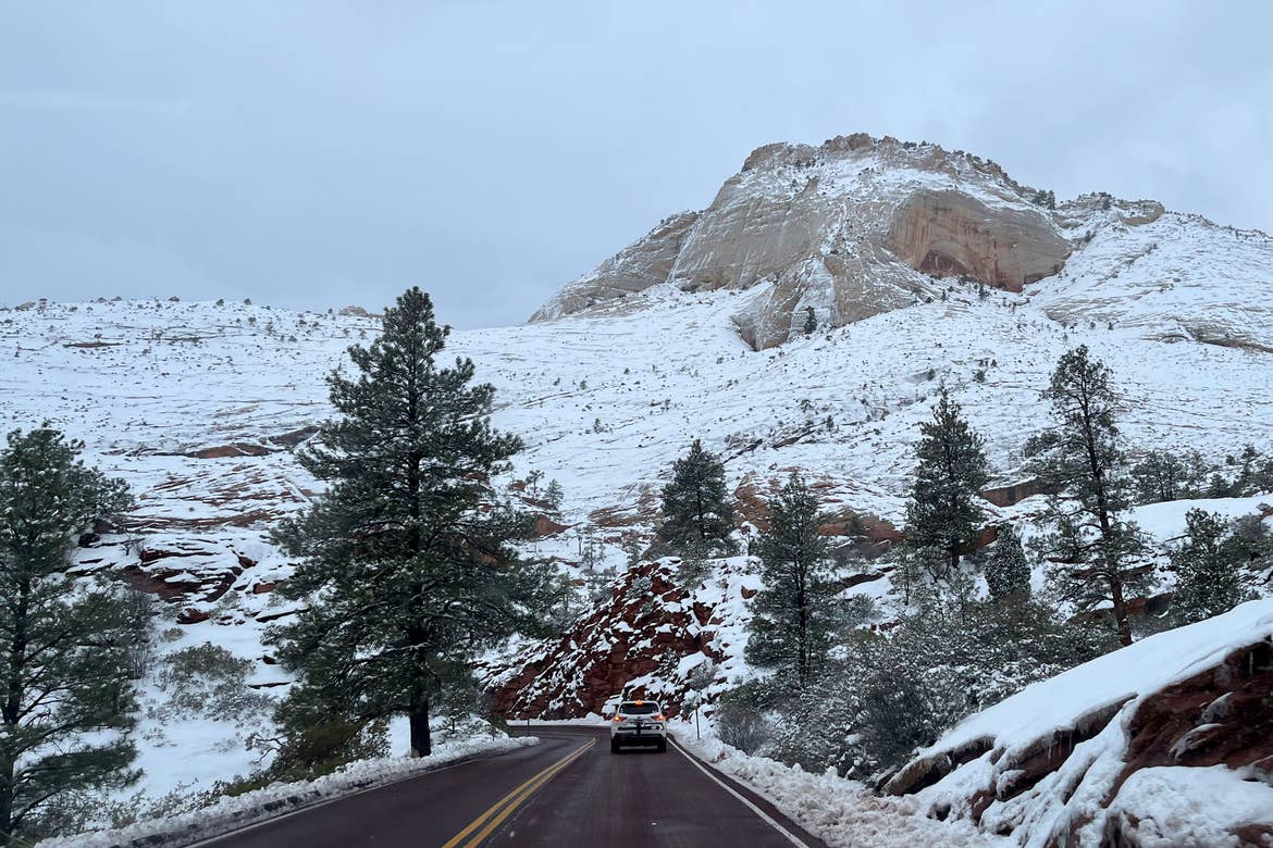 A snow-covered red rock formation looms above a road with a silver suv driving straight ahead. 