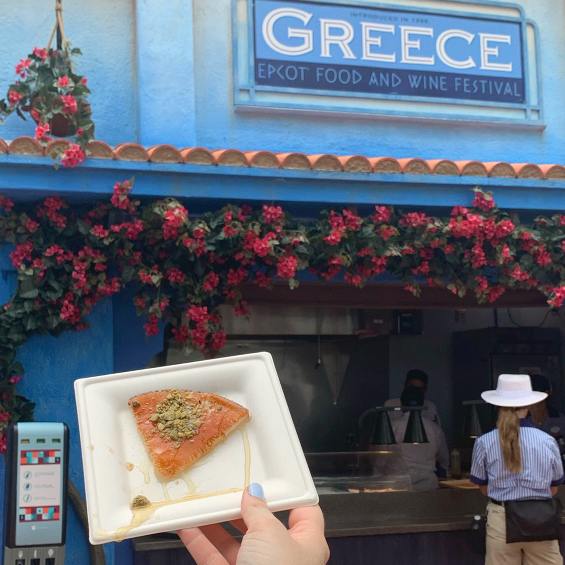 A hand holds a white, square plate containing a Griddled Cheese slice with Pistachios and Honey at the Greece kiosk at Epcot.