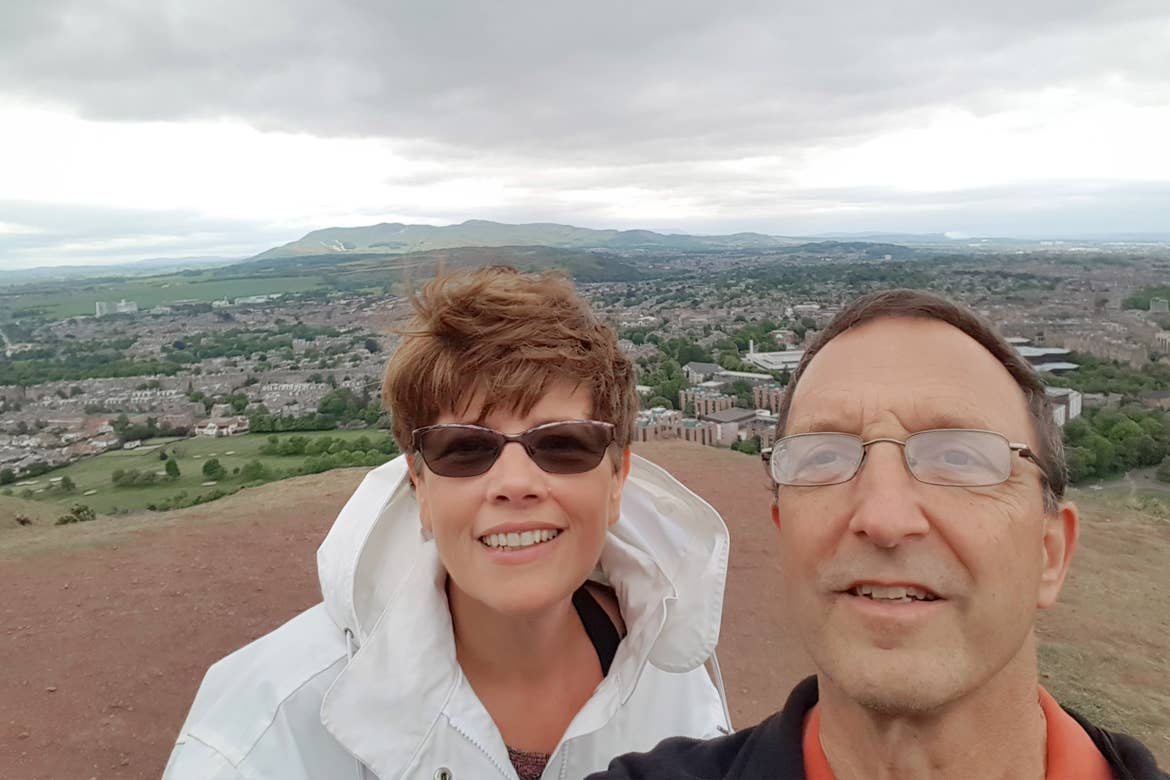 A caucasian male (right) wearing a navy windbreaker and a caucasian woman wearing a white windbreaker jacket stand on a clay hill overlooking Arthur's Seat in Edinburgh, Scotland.