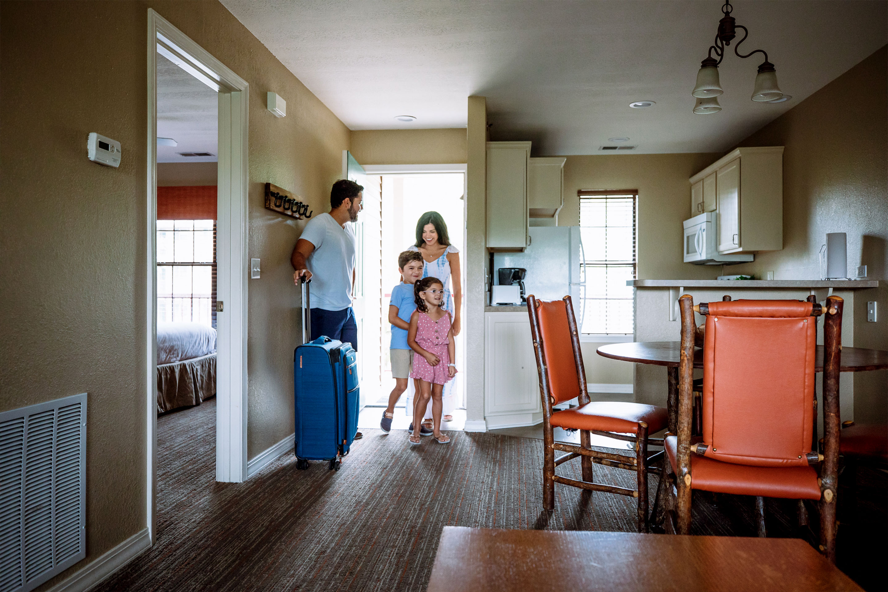 A man (left), woman (back-right), a young boy (middle-back) and girl (front) walk into a villa while pulling suitcases.