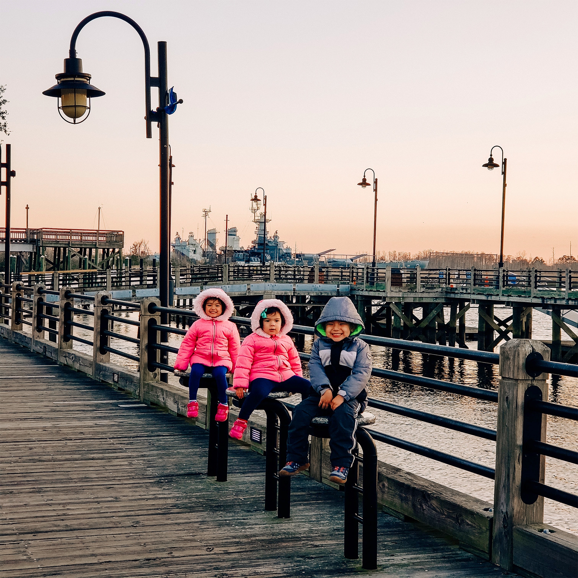 Three Asian Pacific Islander toddlers (left to right: Two girls and a boy) wear pink and blue winter coats while seated on safety railing near a dock for battleships.