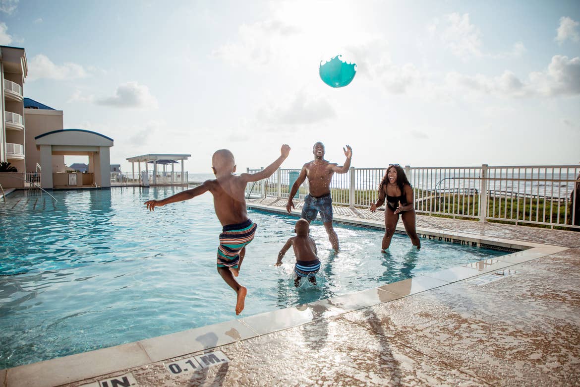 A woman, man and two young boys jump into an infinity pool near a beachfront with a beach ball.
