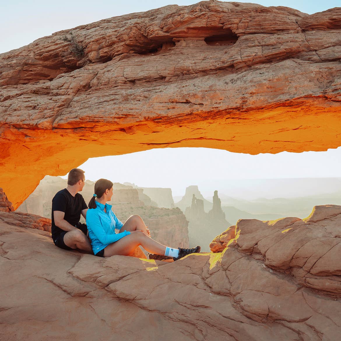 A man in black t-shirt and shorts sits behind a woman in blue pullover and shorts sitting on a red rock formation overlooking the Moab rock formations.