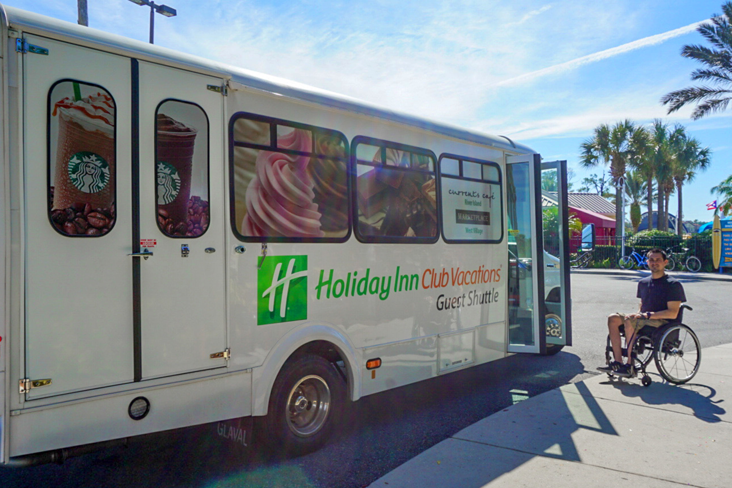 Author, Danny Pitaluga (right) sits in his wheelchair wearing a black shirt and khaki shorts next to a white hotel shuttle bus that reads 'Holiday Inn Club Vacations.'
