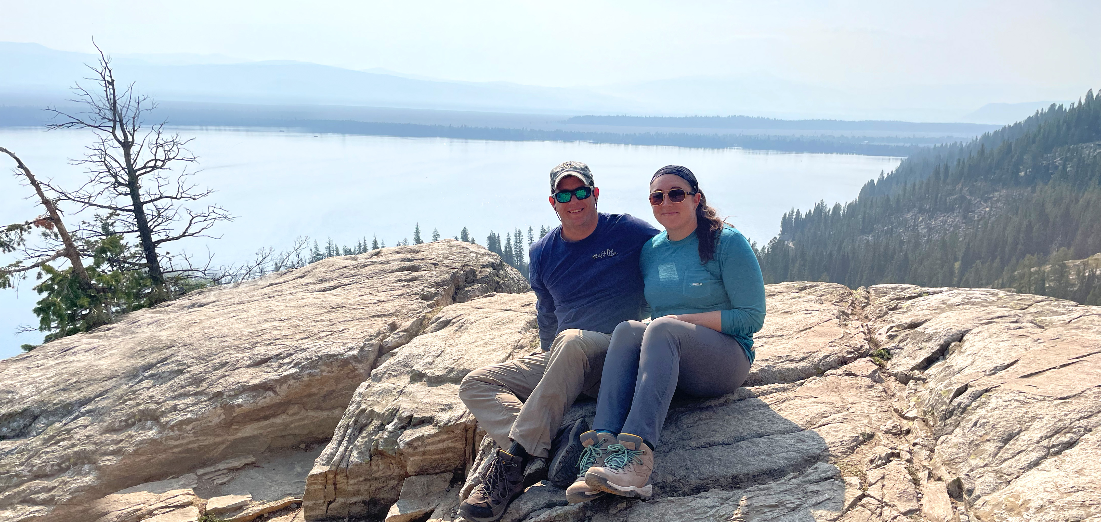 A woman and man in a blue, long sleeve shirt, grey pants, and brown hiking boots sits on a rock formation on a mountain overlooking a lake.