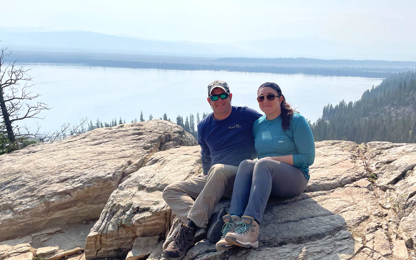 A woman and man in a blue, long sleeve shirt, grey pants, and brown hiking boots sits on a rock formation on a mountain overlooking a lake.