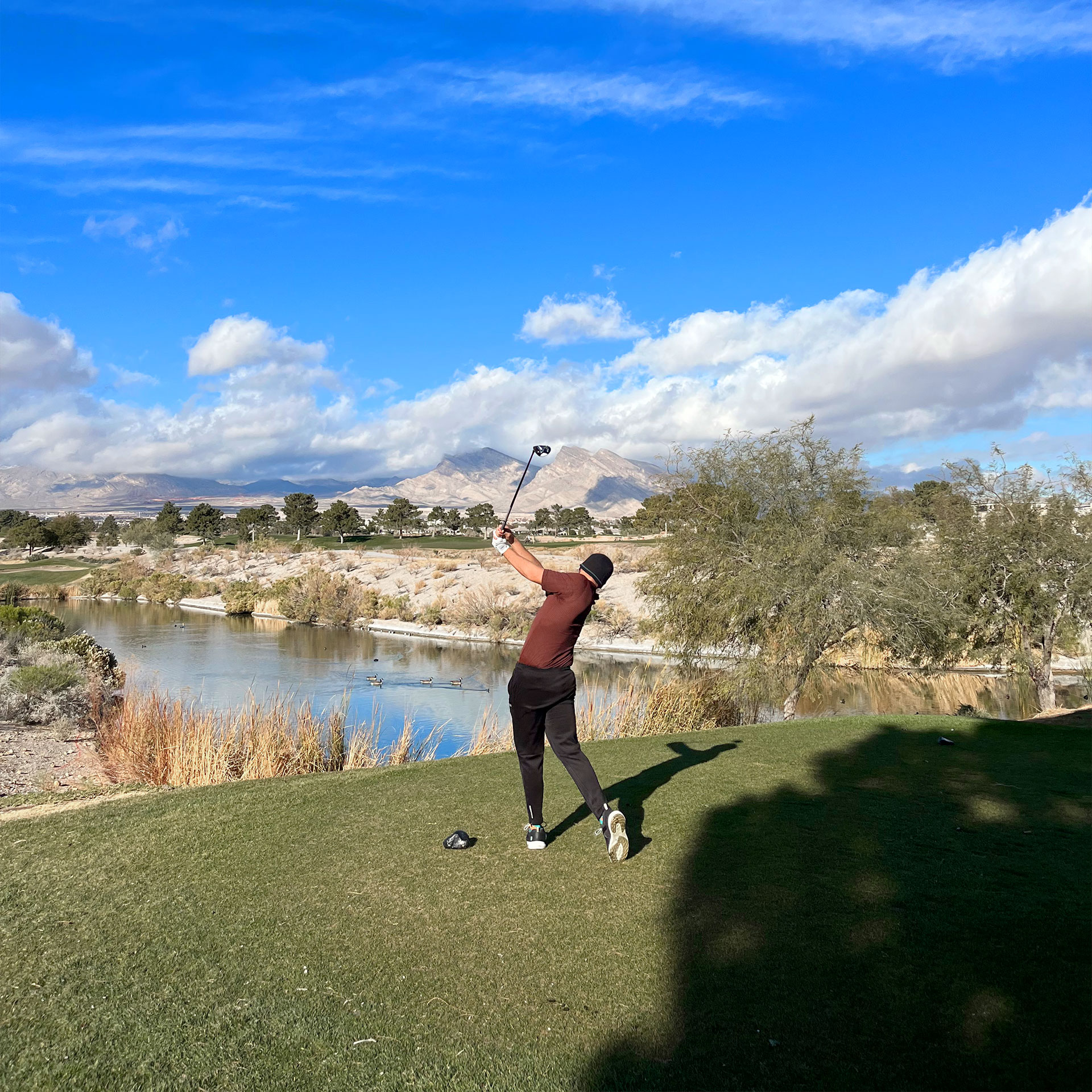 A man in a maroon shirt, black pants and black knitted cap uses a driver club on a golf course with a blue sky and snow-capped mountain range in the distance.
