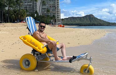 Featured Contributor, Danny Pitaluga, wears sunglasses and swim trunks while sitting in a yellow beach wheelchair on the sand as waves roll in.