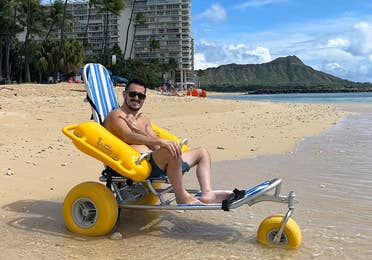 Featured Contributor, Danny Pitaluga, wears sunglasses and swim trunks while sitting in a yellow beach wheelchair on the sand as waves roll in.