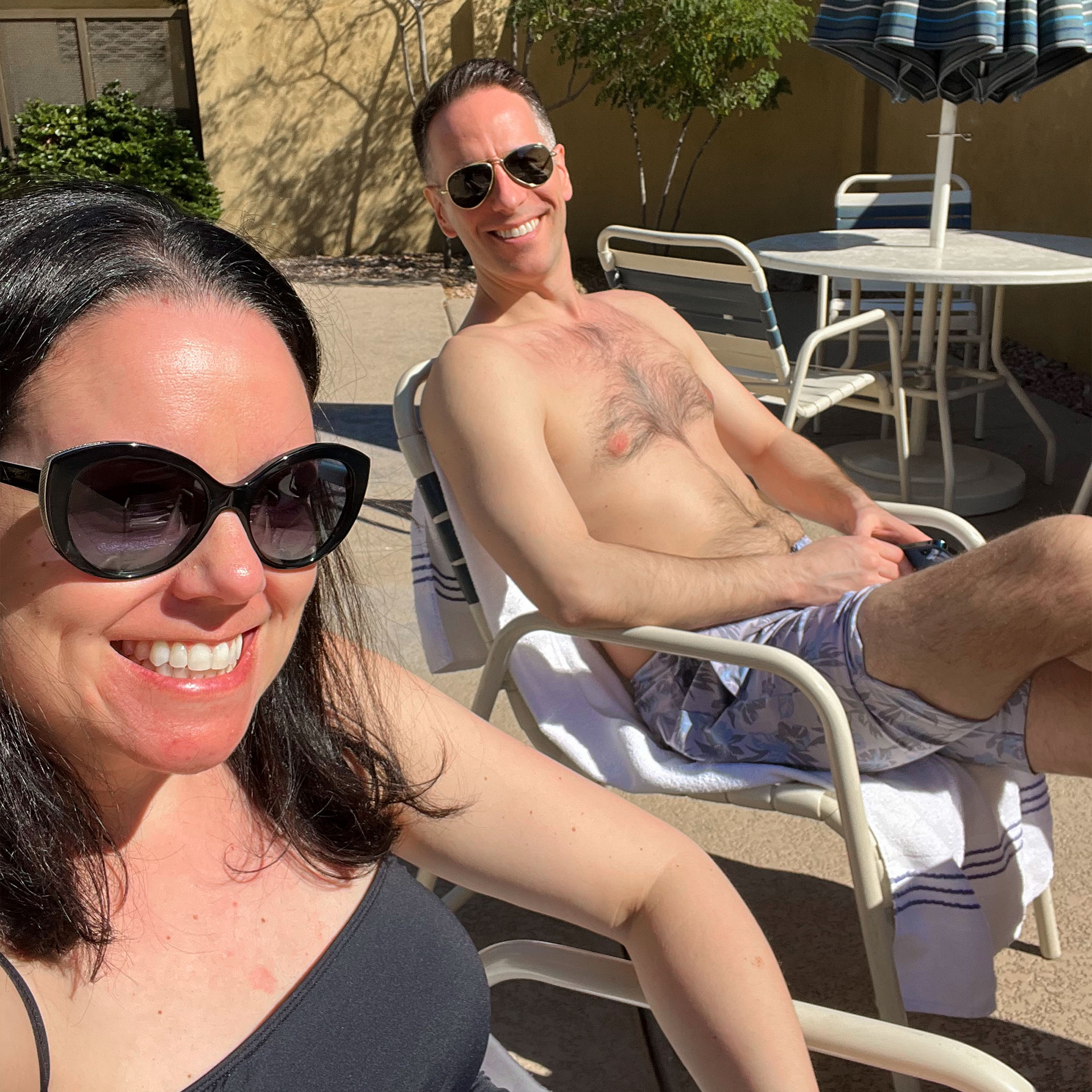 A woman in black swimwear and sunglasses (left) and a man in swim trunks and sunglasses (right) sit on poolside patio chairs.