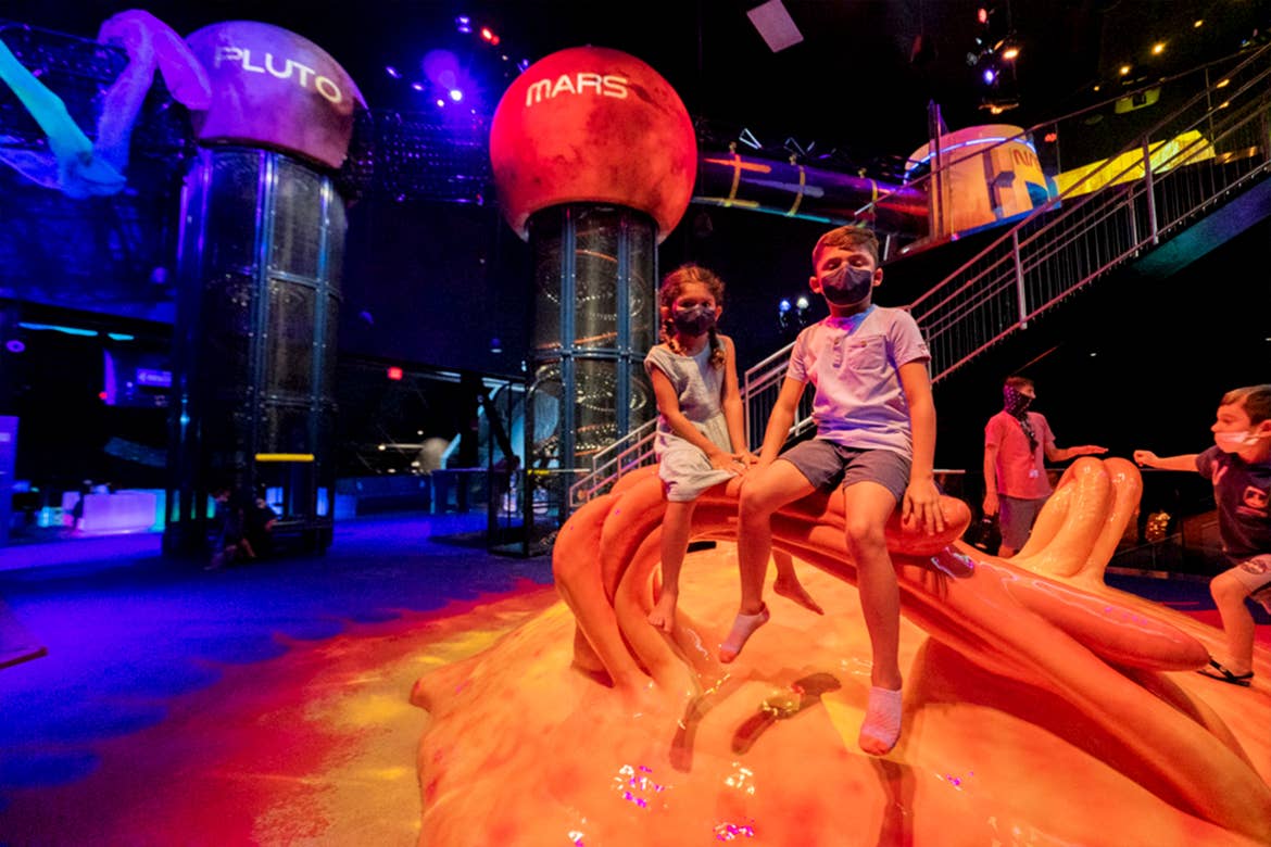 A young boy wearing a blue shirt, shorts and black face mask sits on a scaled structure next to a young girl in a blue dress and black face mask.