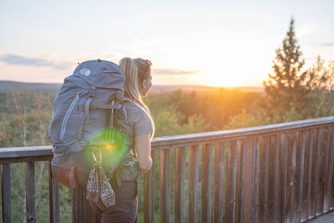 Featured Contributor, Ashlyn George, wears a backpack and watches the sunset above pine trees.