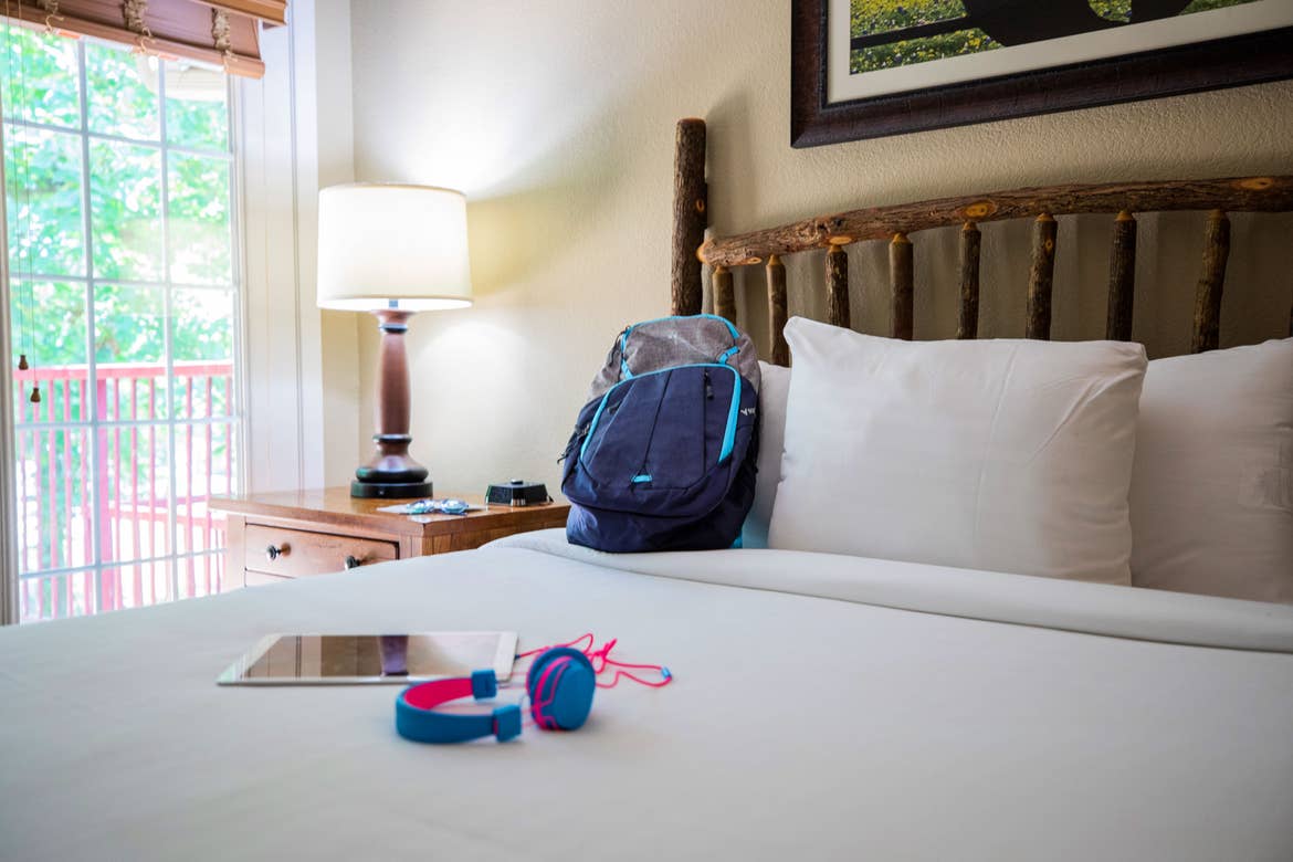 A pair of headphones, an iPad and backpack are placed on a bed with white linens.