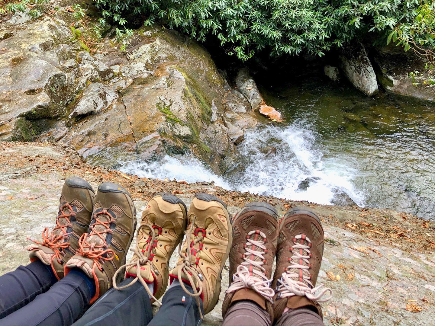 Featured Contributor, Jennifer C. Harmon (left) and her two girlfriends wear hiking boots as their feet sit near the top of a waterfall.
