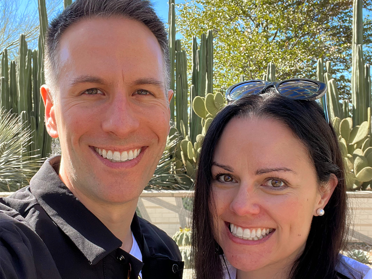 A man in a black jacket (left) and a woman in a white jacket and sunglasses (right) stand in front of cacti outdoors.