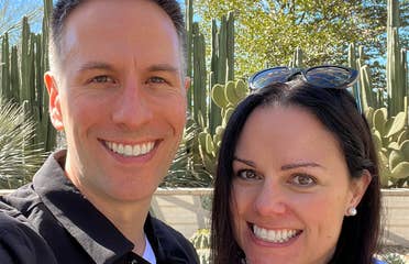 A man in a black jacket (left) and a woman in a white jacket and sunglasses (right) stand in front of cacti outdoors.