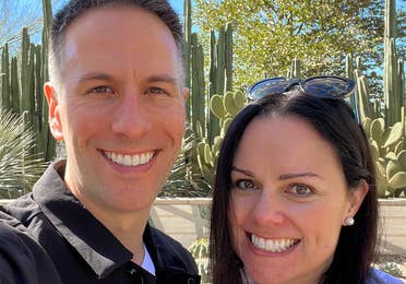 A man in a black jacket (left) and a woman in a white jacket and sunglasses (right) stand in front of cacti outdoors.