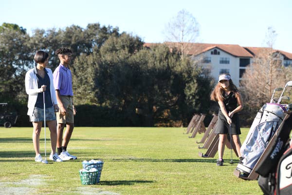 Girl holding a golf club at the driving range as two people watch.