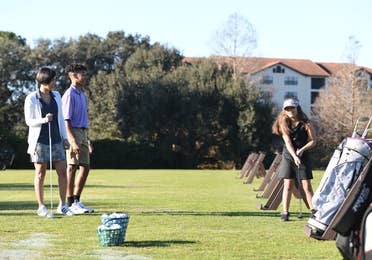 Girl holding a golf club at the driving range as two people watch.