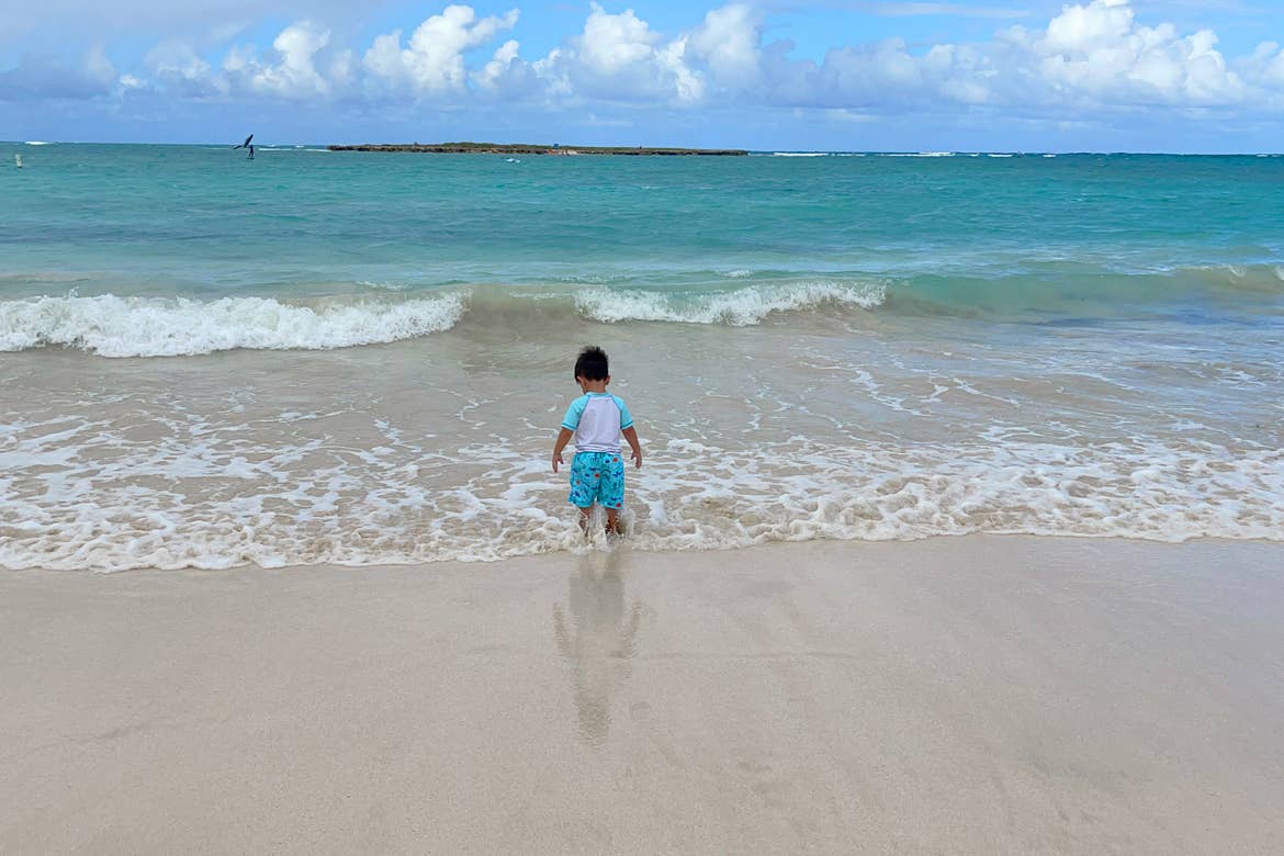 Featured Contributor, Danny Pitaluga's son, Joey, stands in the sand on the beach in swimwear as waves roll in.
