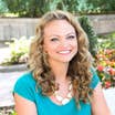 Featured Contributor, Angie Orth of @angieaway, sits outdoors near a garden wearing a necklace with white beads and a blue tee shirt.