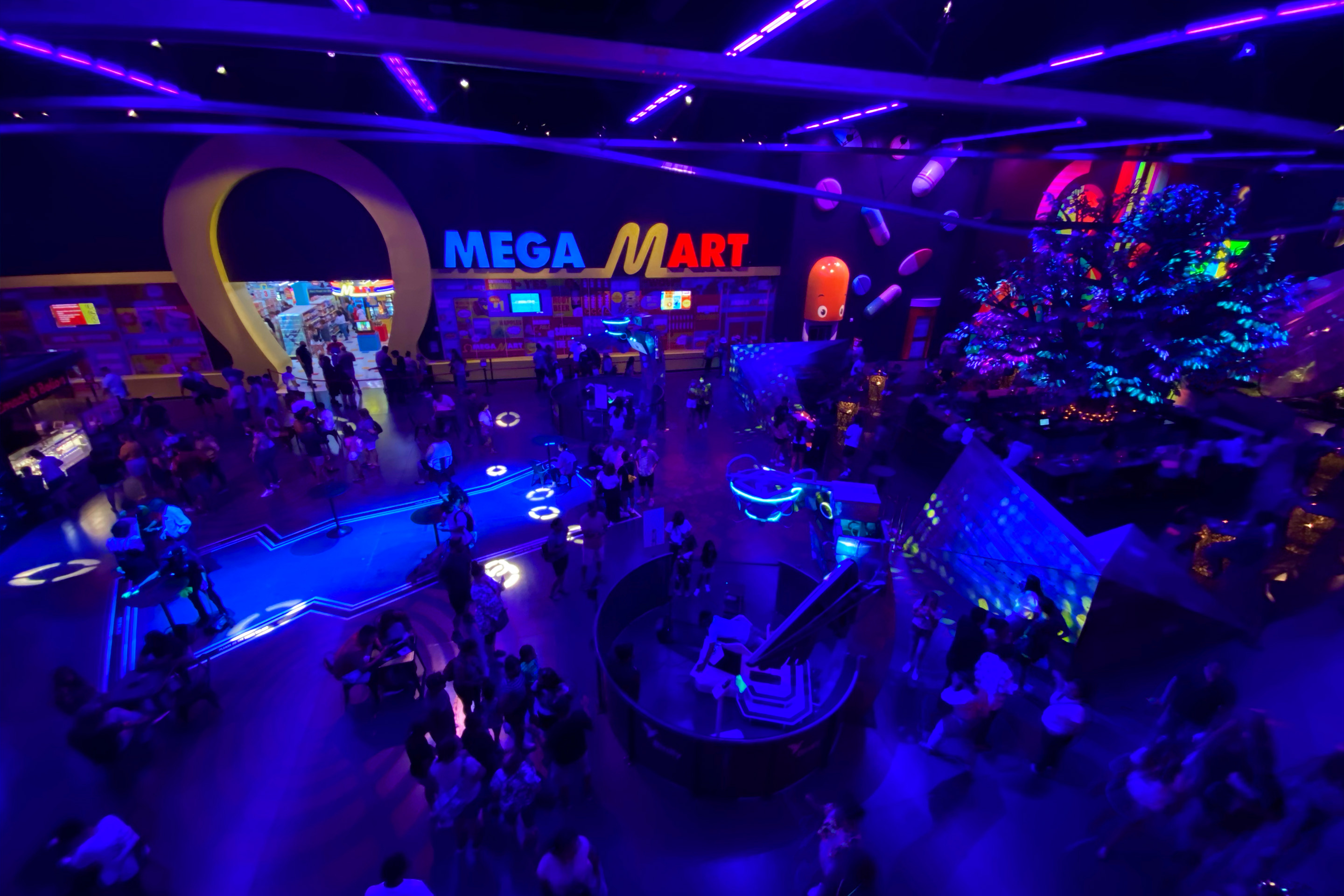 An interior space with blue lighting and an illuminated marquee that reads, 'Mega Mart' in yellow and red neon contains numerous guests.