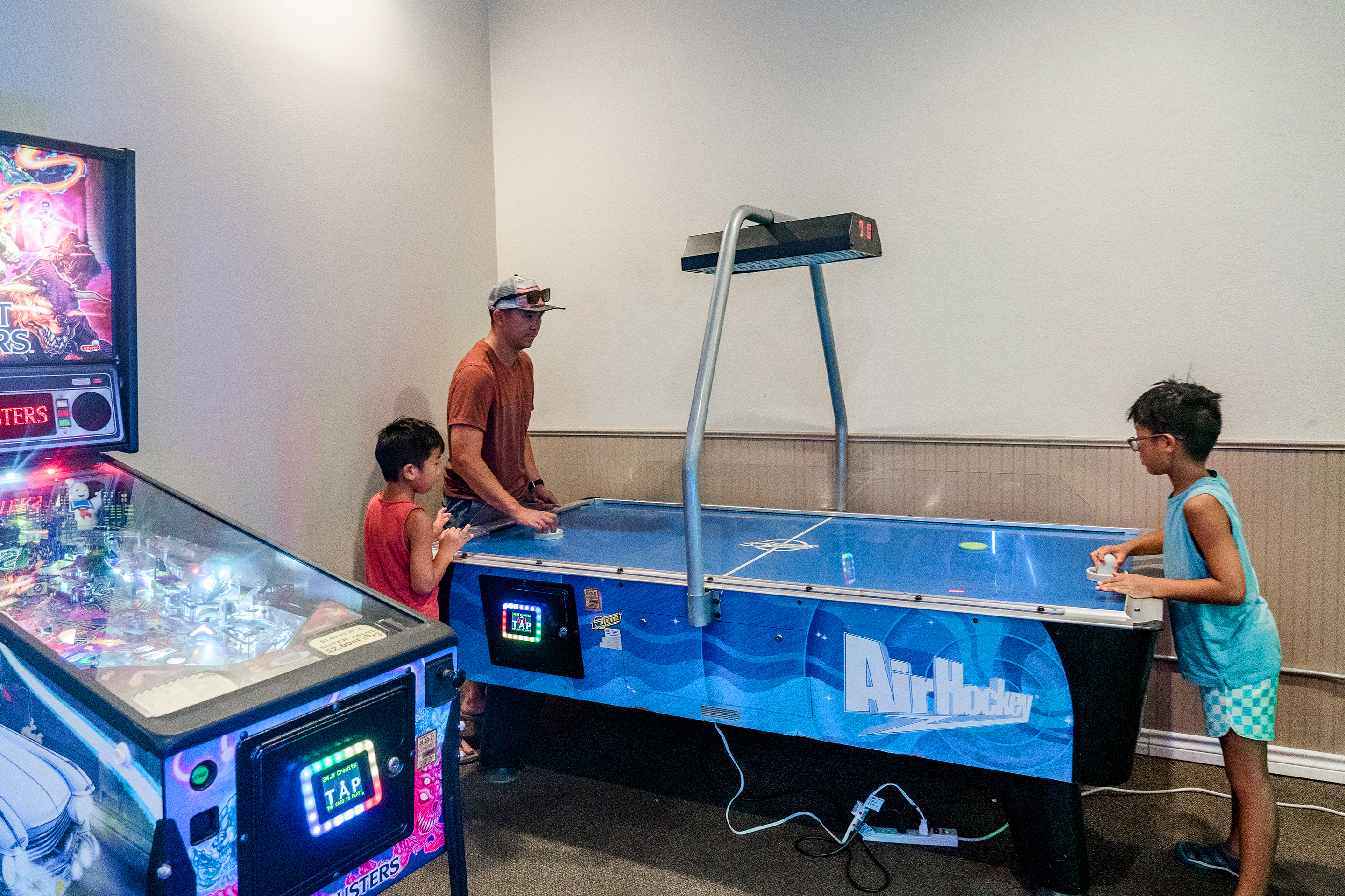 An Asian man (back-left) and two young Asian boys (left and right) play at a blue air hockey table near an illuminated pinball machine at our Villages resort in Flint, Texas.