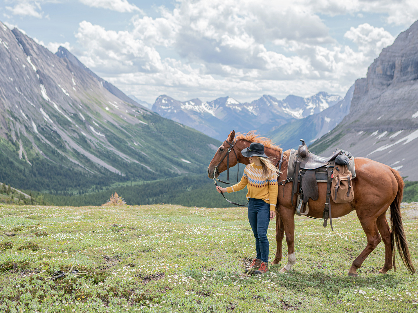 Featured Contributor, Ashlyn George, stands with a horse in front of the Canadian mountain range.